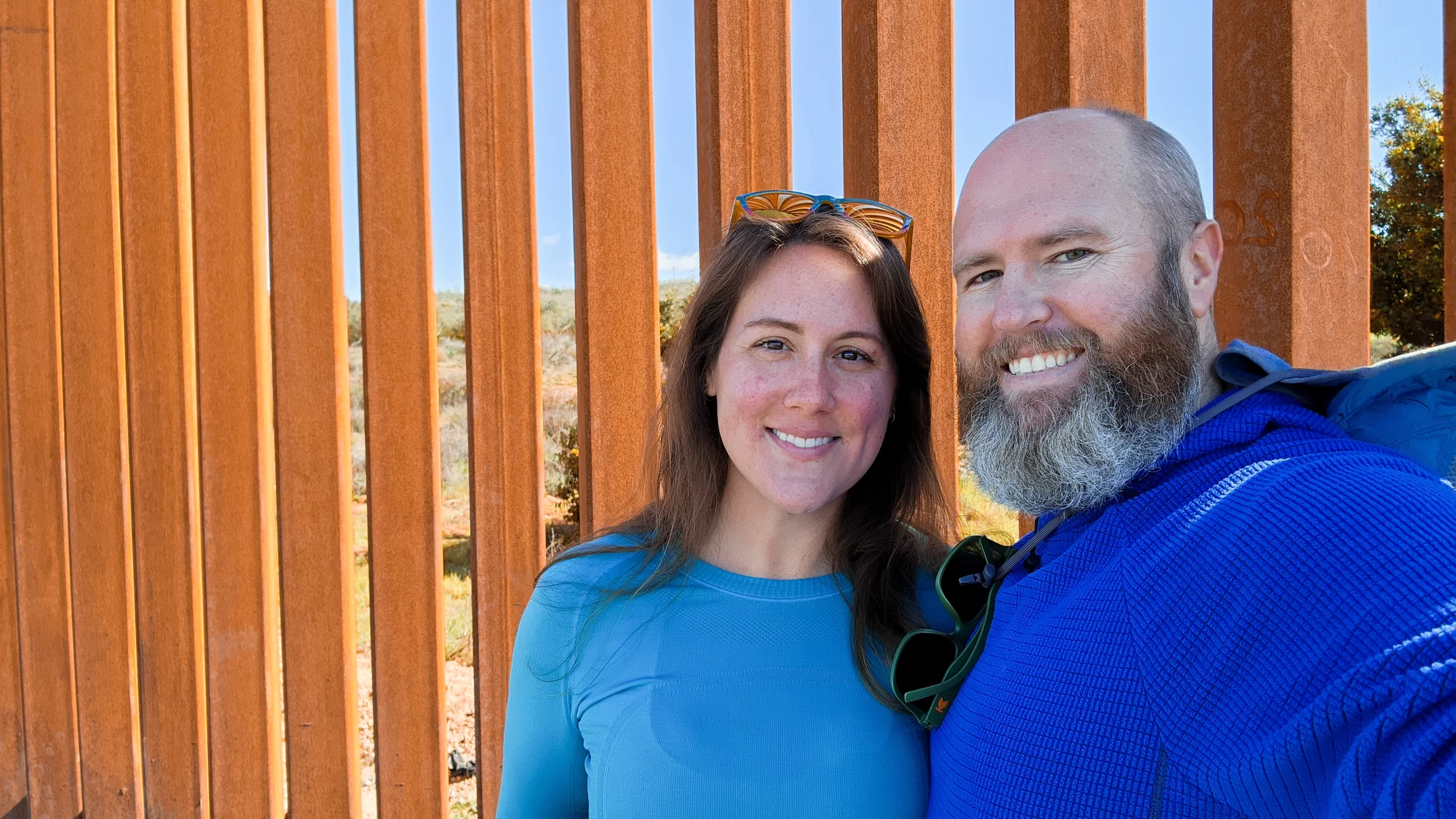 Tom and Elyse smiling together before the US-Mexico patinated steel border wall.