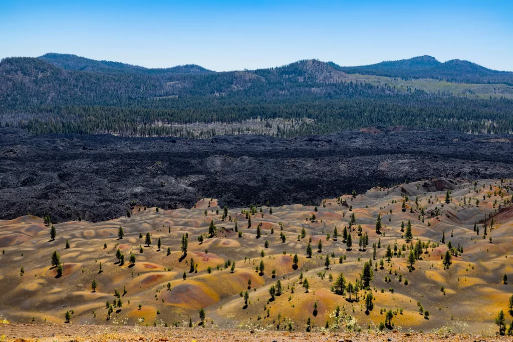 Towering forested hills followed by the black rough Fantastic Lava Beds, and the colorful Painted Dunes in the foreground.